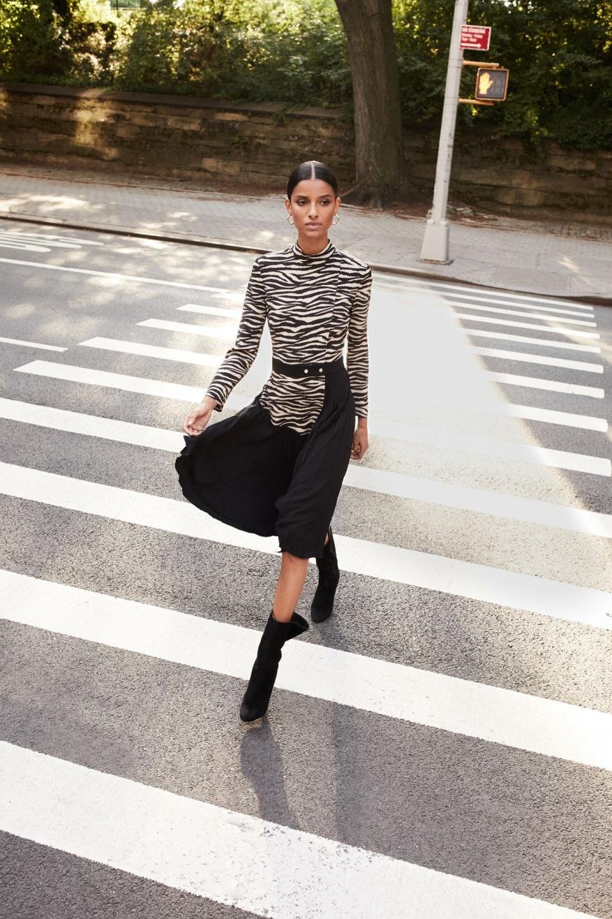 Elegant woman in zebra-print top and black skirt strides confidently across a crosswalk on a sunny city street, fashionably chic.