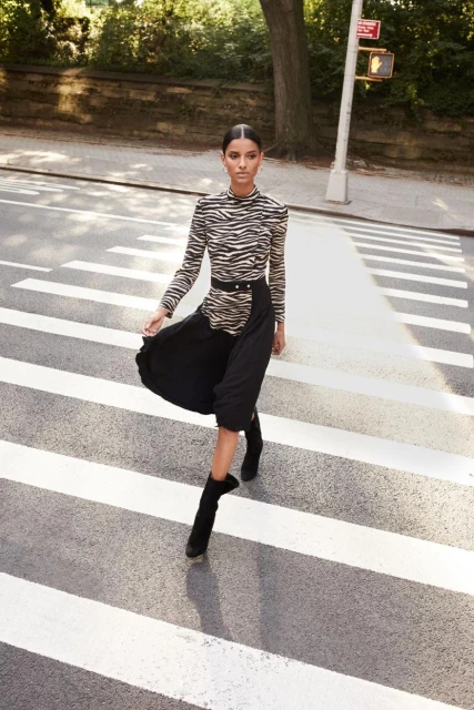 Elegant woman in zebra-print top and black skirt strides confidently across a crosswalk on a sunny city street, fashionably chic.