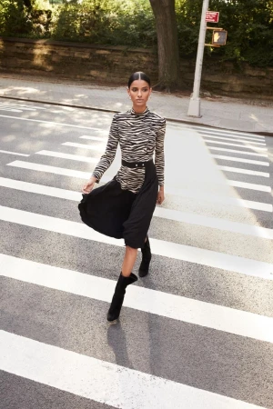 Elegant woman in zebra-print top and black skirt strides confidently across a crosswalk on a sunny city street, fashionably chic.