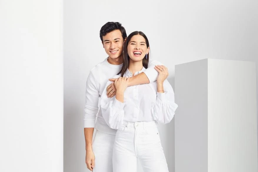 Smiling couple embraces indoors in a bright photo studio. The pair is dressed in white and posed in front of white walls and props.