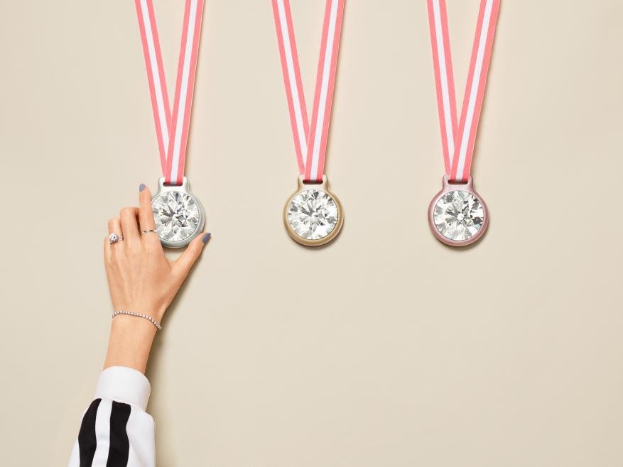 Three hanging diamond pendants on pink ribbons, with a woman's arm holding the leftmost pendant. Cream-colored background. Jewelry concept.