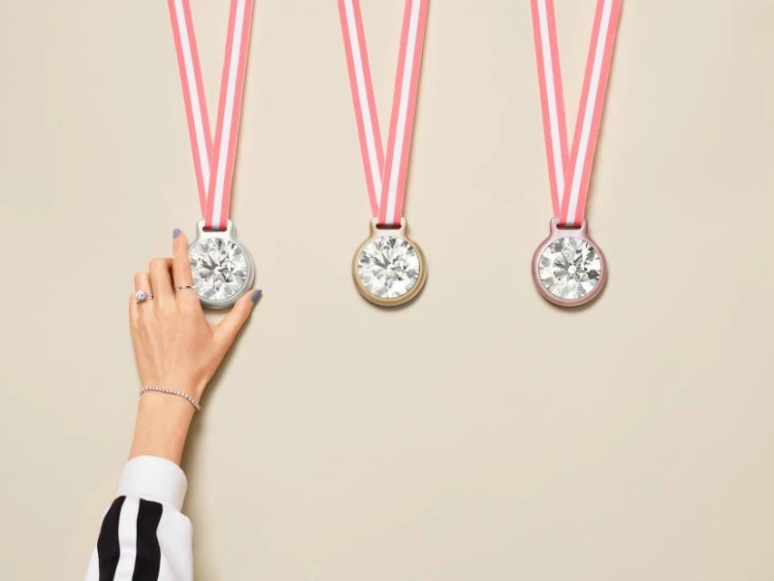 Three hanging diamond pendants on pink ribbons, with a woman's arm holding the leftmost pendant. Cream-colored background. Jewelry concept.