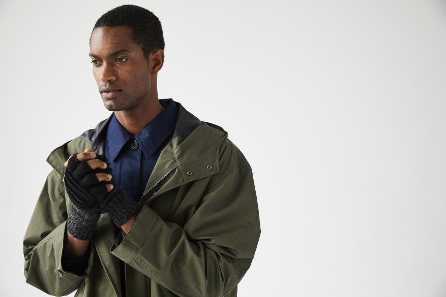 An adult male in an olive green jacket and fingerless gloves in a studio. He is looking left with hands clasped together.
