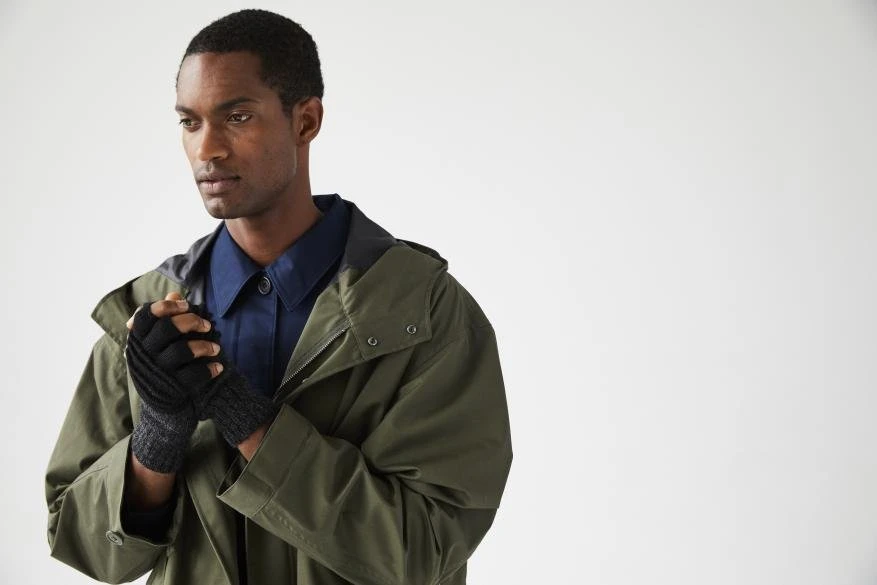 An adult male in an olive green jacket and fingerless gloves in a studio. He is looking left with hands clasped together.