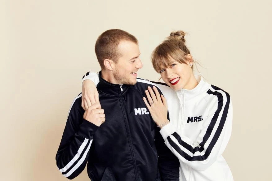 Smiling couple in matching black and white tracksuits with 'Mr.' and 'Mrs.' on them, posing against a cream-colored background in a studio setting.