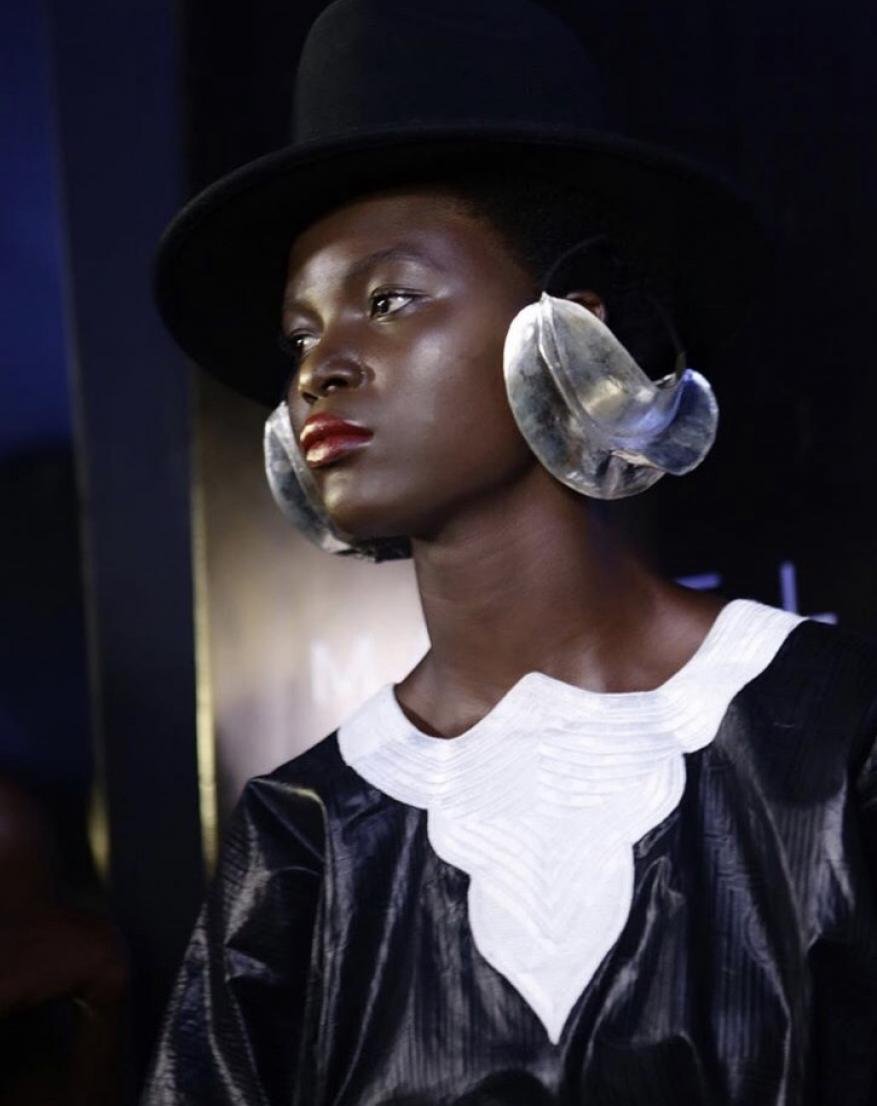 Portrait of Black woman with large transparent earrings and a hat. She is wearing a black top with white decorative collar.