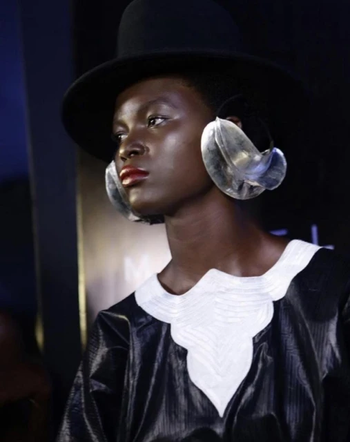 Portrait of Black woman with large transparent earrings and a hat. She is wearing a black top with white decorative collar.