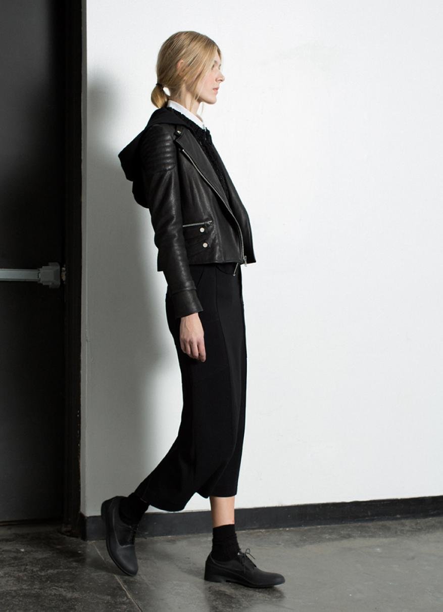 Full length shot of a slender woman with blonde hair in a ponytail wearing a black leather jacket, black culottes and matching shoes.