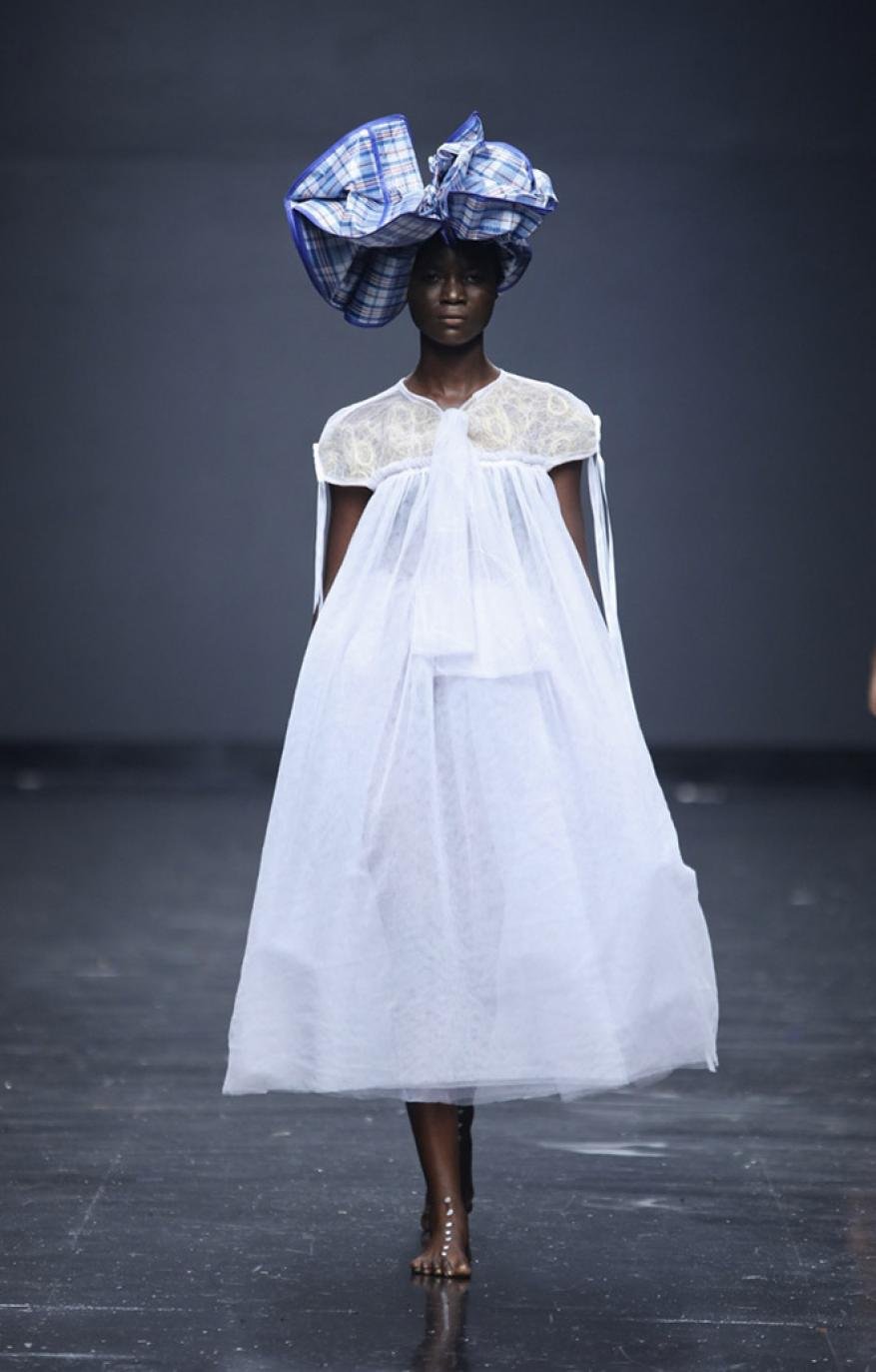 An adult female model showcases a flowing white dress and oversized blue-and-white patterned hat on a fashion runway with a dark gray background.