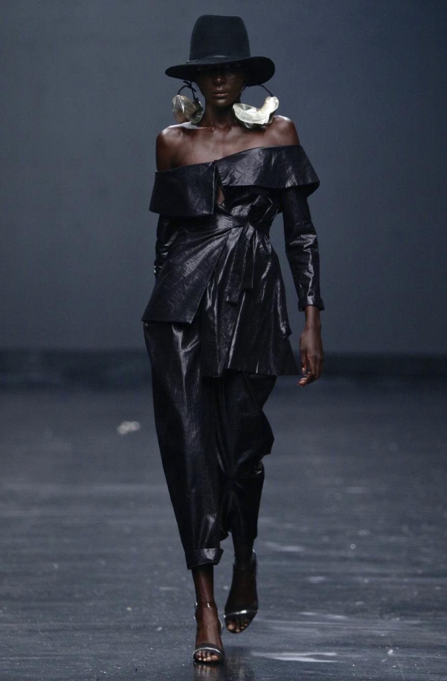Runway shot of a Black fashion model wearing a shiny black off-shoulder outfit and large disc earrings against a dark studio backdrop.