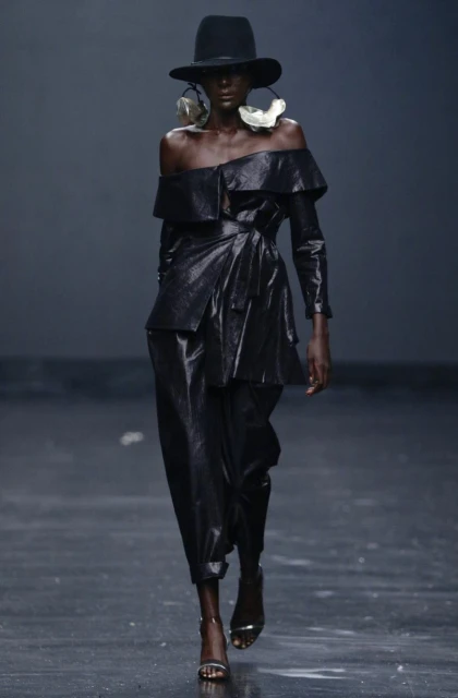 Runway shot of a Black fashion model wearing a shiny black off-shoulder outfit and large disc earrings against a dark studio backdrop.