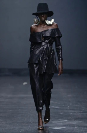 Runway shot of a Black fashion model wearing a shiny black off-shoulder outfit and large disc earrings against a dark studio backdrop.