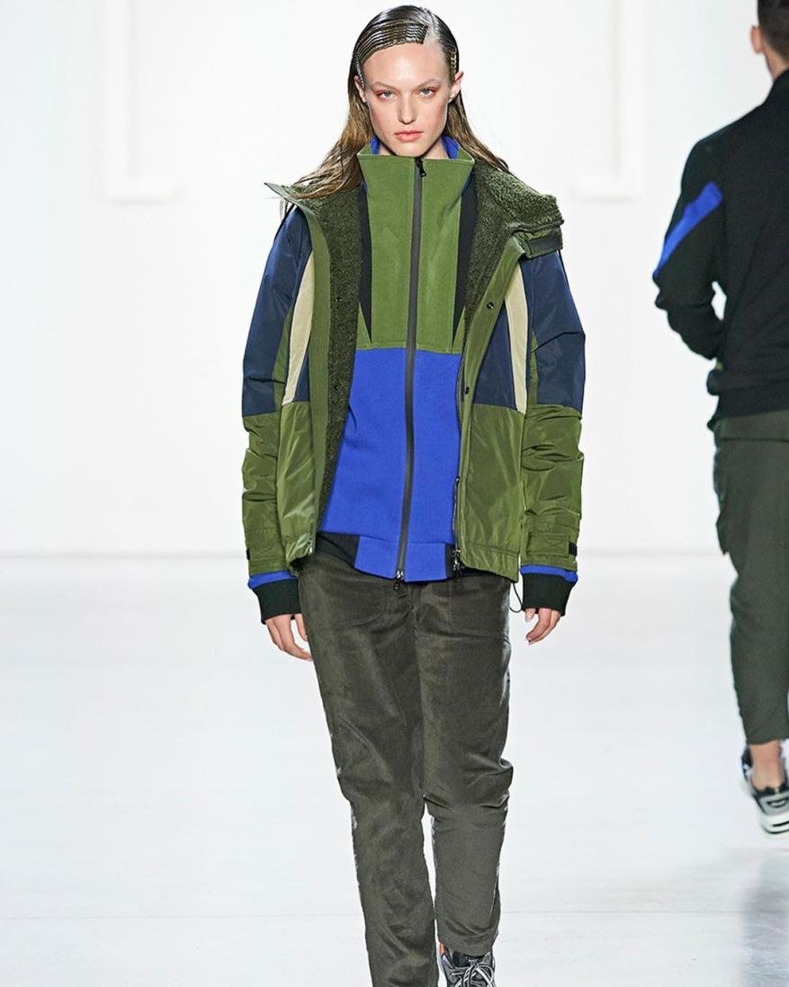 A fashion model walks the runway wearing a color-blocked jacket, green pants, and sneakers in a brightly lit indoor fashion show.