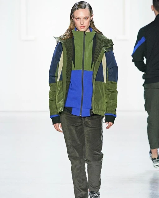 A fashion model walks the runway wearing a color-blocked jacket, green pants, and sneakers in a brightly lit indoor fashion show.