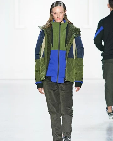 A fashion model walks the runway wearing a color-blocked jacket, green pants, and sneakers in a brightly lit indoor fashion show.