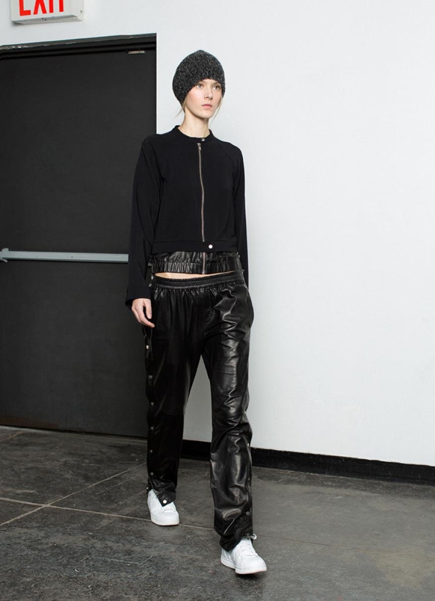 An adult female fashion model in a black top and shiny black pants poses in a studio with white walls and a dark door.