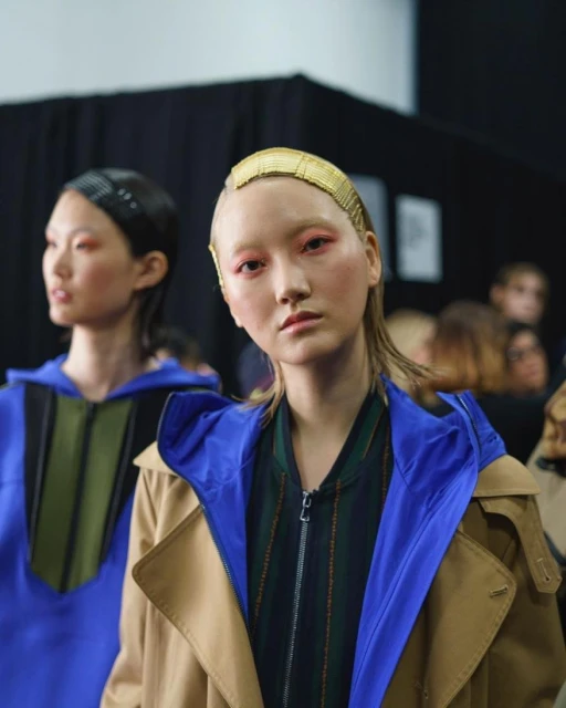 Two asian fashion models pose in a backstage setting, wearing modern clothing and distinctive gold hair accessories, medium shot.