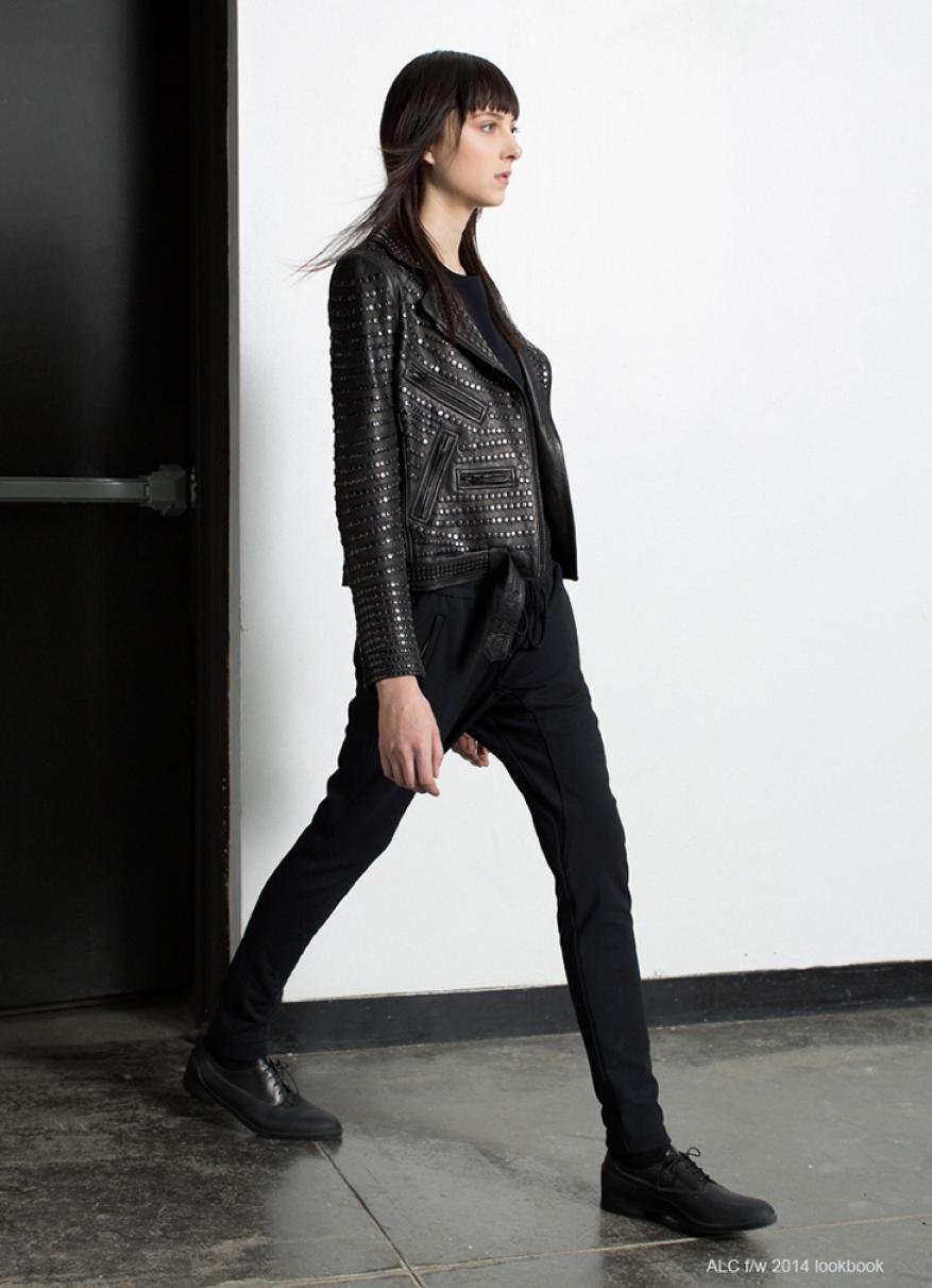 Woman walks in black leather jacket and black pants against a white wall with a black door, showcasing a modern fashion style.