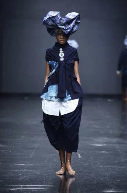 Runway shot of model in blue patterned head wrap and blue tie-dye outfit walking the runway at fashion show with grey backdrop.