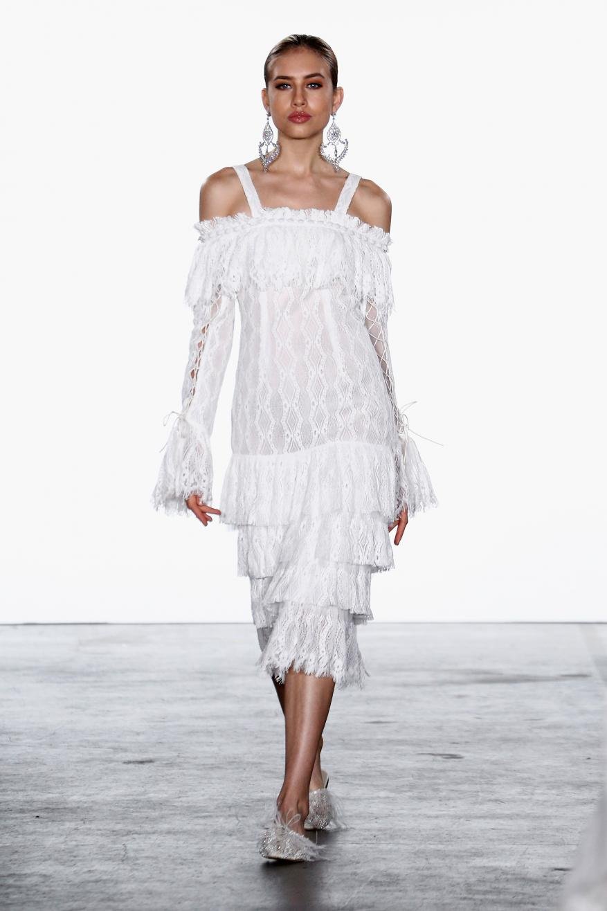 Runway model presents white lace midi dress with ruffles, long sleeves, and statement earrings, during a fashion show against white background.