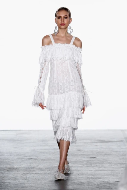 Runway model presents white lace midi dress with ruffles, long sleeves, and statement earrings, during a fashion show against white background.