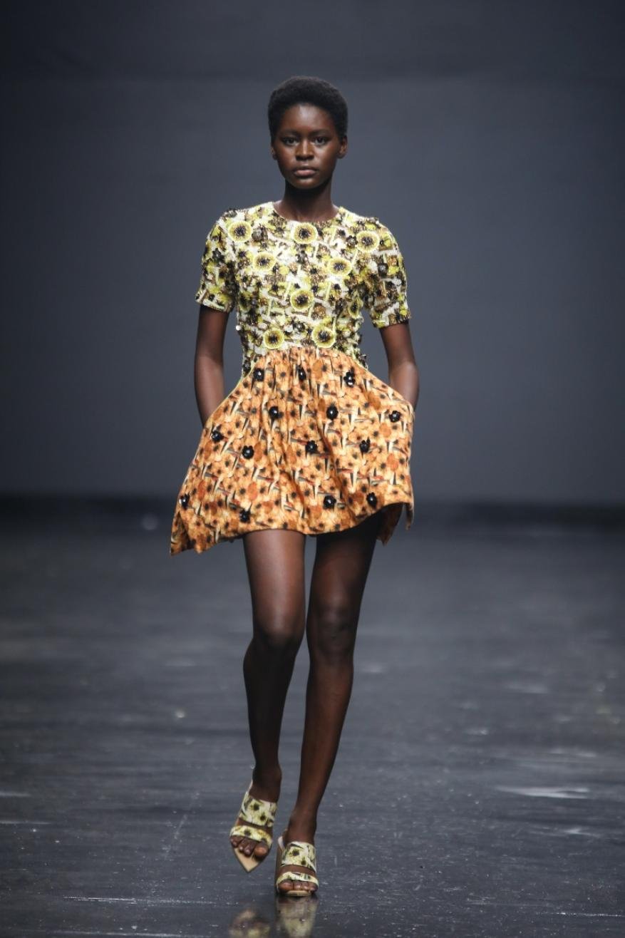 Runway fashion show featuring an African woman, wearing a patterned mini-dress and heels, walking confidently against a dark gray background.