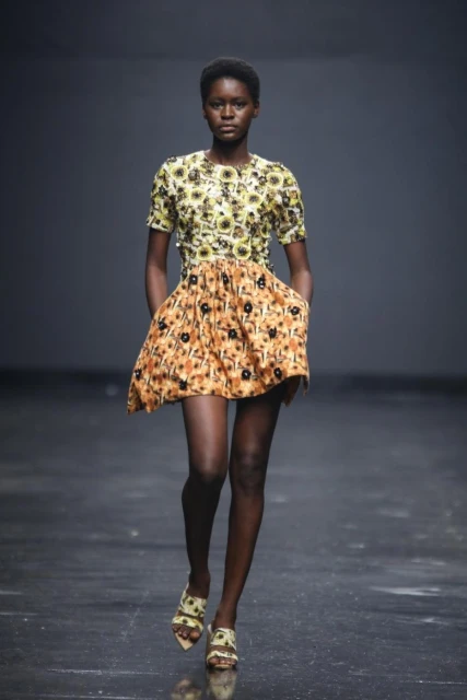 Runway fashion show featuring an African woman, wearing a patterned mini-dress and heels, walking confidently against a dark gray background.