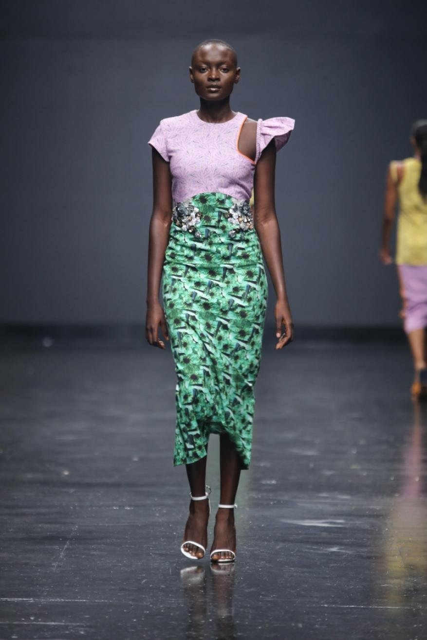 Runway fashion show featuring a black female model in a lavender and green patterned dress with white strappy high heels on a dark stage.