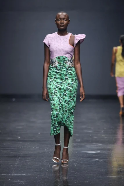 Runway fashion show featuring a black female model in a lavender and green patterned dress with white strappy high heels on a dark stage.