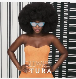 Black woman with large afro wearing orange strapless top and turquoise cat-eye sunglasses against patterned background