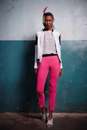 Model wearing a white jacket and pink trousers with a feather hairpiece standing before a weathered teal and white wall