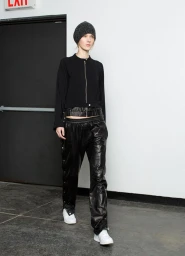 Model wearing black beanie, black zip jacket, and shiny black pants walking indoors against a white wall.