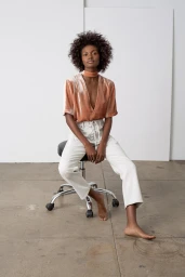 Black woman with curly hair seated on a rolling stool, wearing a peach velvet top and white lace-up pants.