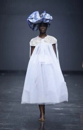 Black model walking runway in sheer white dress and sculptural blue patterned headpiece styled by Joan Reidy