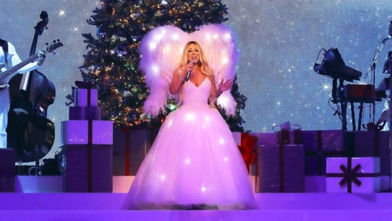 Adult female singer in a white, light-up gown and wings on stage at a holiday music concert with musicians and Christmas tree backdrop.