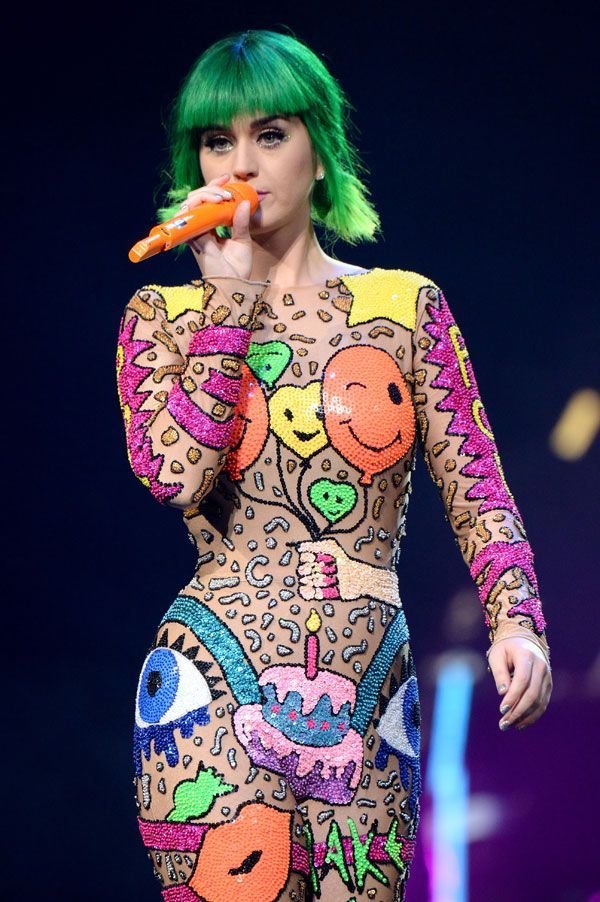 Female musician with green hair performs on stage, singing into a microphone, wearing a graphic bodysuit at a concert.