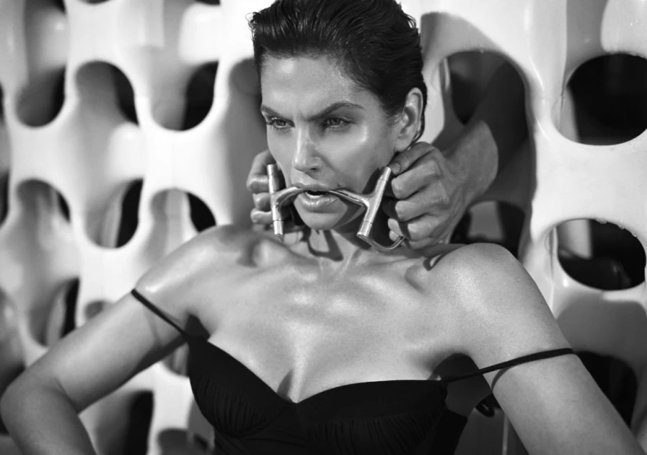 Monochrome portrait of a woman with a metal object in her mouth, hands behind her head, and a patterned backdrop.