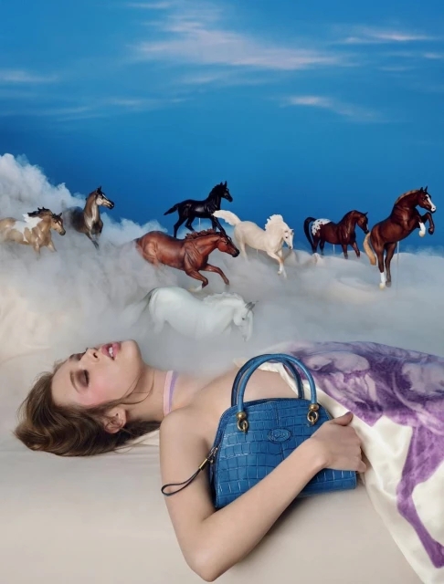 Dreamy fashion photo featuring a young woman lying down with a blue purse and several horses in the background against a blue sky.