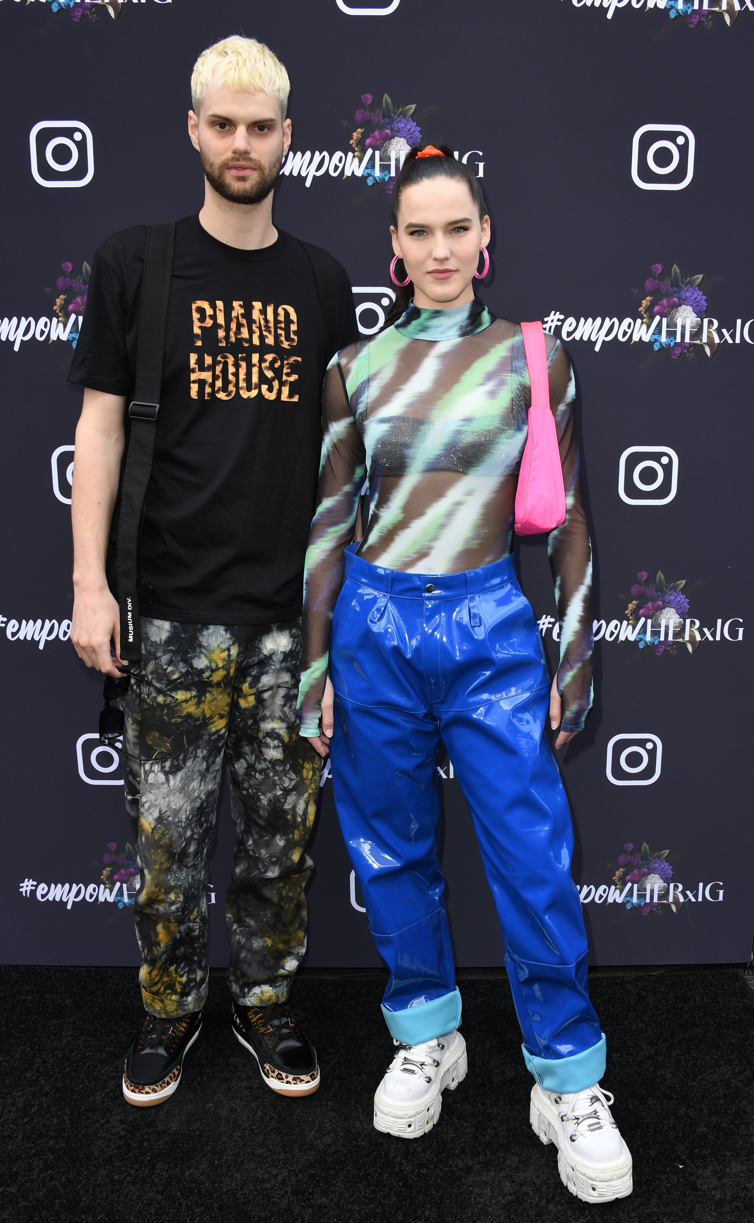 Two people pose in front of a branded backdrop. The male figure wears a graphic t-shirt and patterned pants. The female figure wears a sheer top and blue pants.