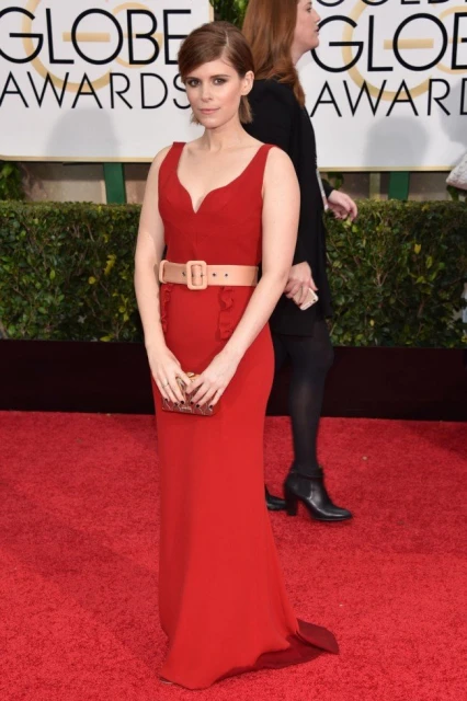 Kate Mara attends an awards show in a red dress with a belt, standing on a red carpet with another person standing in the background.