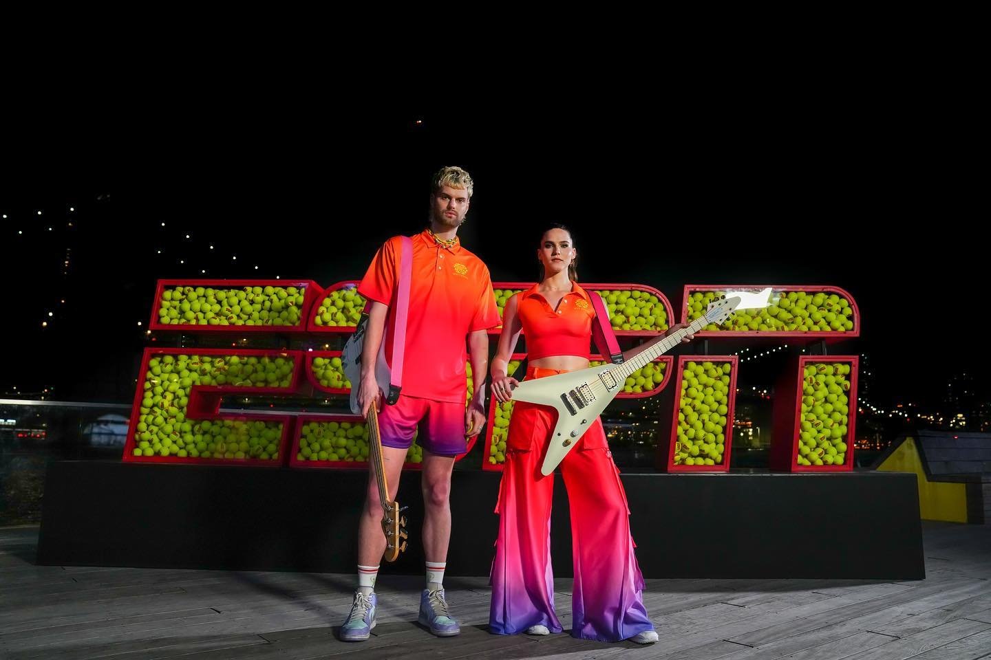 Two fashion models pose in sunset-ombre apparel against an ESPN logo made of tennis balls at an outdoor night event.