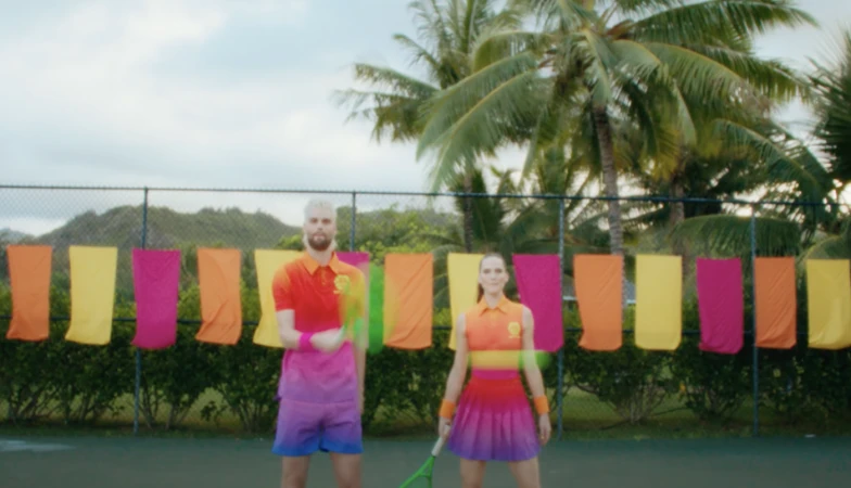 Two tennis players wearing retro-style outfits stand on a green court with hanging colorful towels, palm trees and blue sky in the background.
