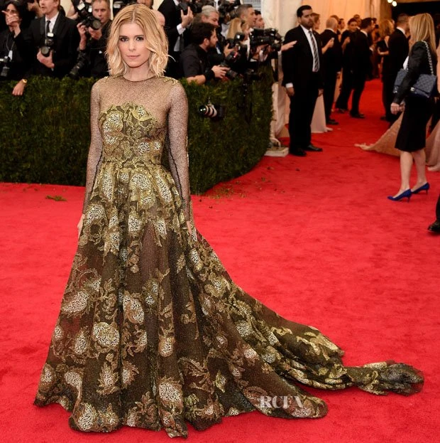 Kate Mara poses on a red carpet in a floral, floor-length gown. She is surrounded by onlookers, photographers, and reporters.