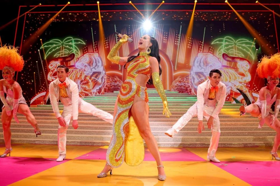 Female singer performs on stage in a shimmering orange and silver dress, backed by dancers in white suits on a brightly lit stage.
