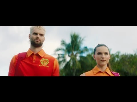 Two people in orange shirts stand outdoors against a backdrop of trees and sky, facing the camera, wearing casual attire and slight smiles.