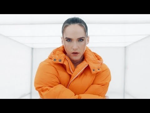 Portrait of a young adult woman with neutral expression in a bright orange puffer jacket, set against a stark white indoor background.