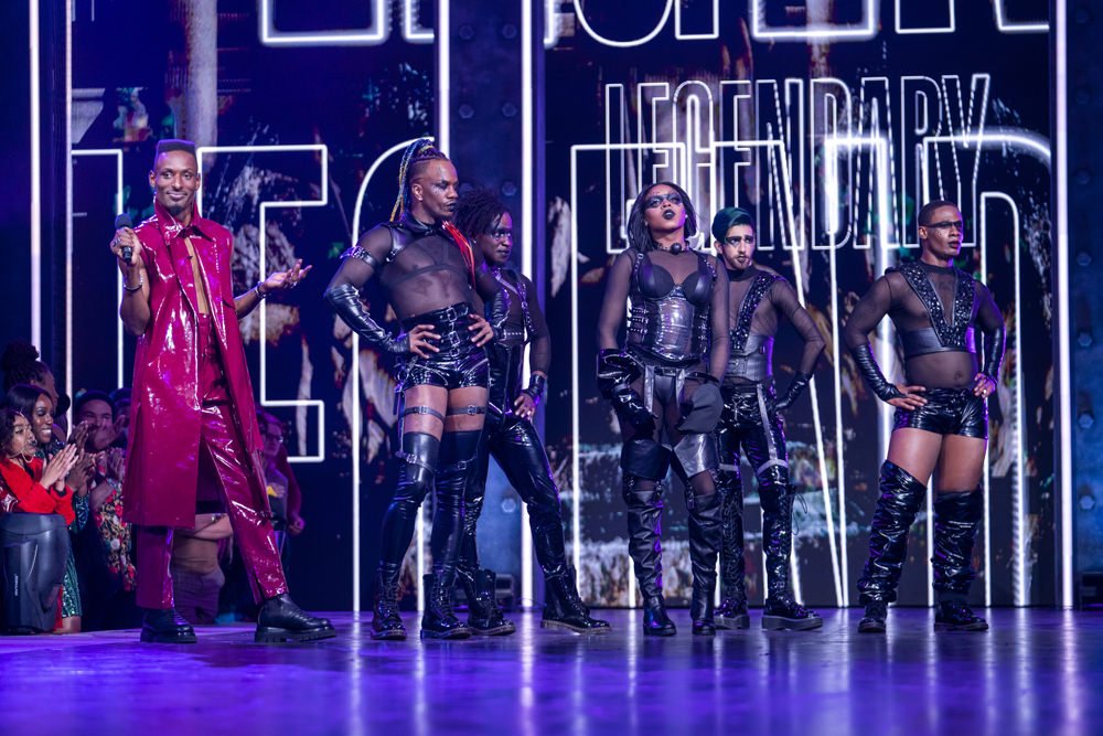 Performance on stage with six fashionably dressed performers in shiny textured clothing in front of a Legendary branded display.