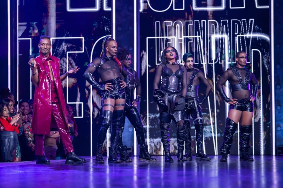 Performance on stage with six fashionably dressed performers in shiny textured clothing in front of a Legendary branded display.