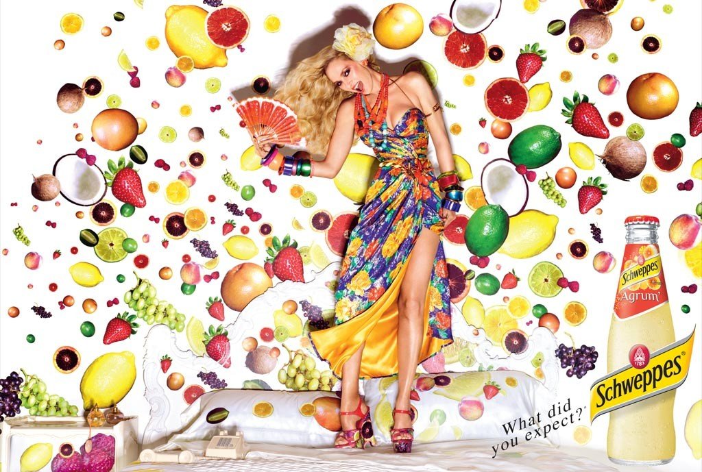 Blond model in floral dress poses in an indoor scene filled with colorful fruits, wearing jewelry, standing on a bed, and holding a fan.