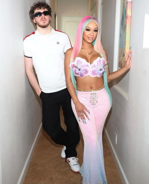 Jack Harlow poses with Saweetie in a hallway; he wears sunglasses, a white shirt, and black pants, while she has multicolored hair and stylish clothes.